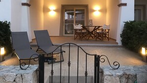 Une terrasse extérieure avec deux chaises longues, une table et des chaises, située sous une zone abritée et éclairée par deux lampes fixées au mur.
