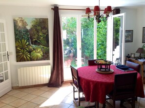 A dining area with a red tablecloth, a painting of tropical foliage, and a view of greenery through the glass door. - Town house in Chevreuse valley (Les Essarts-Le-Roi)
