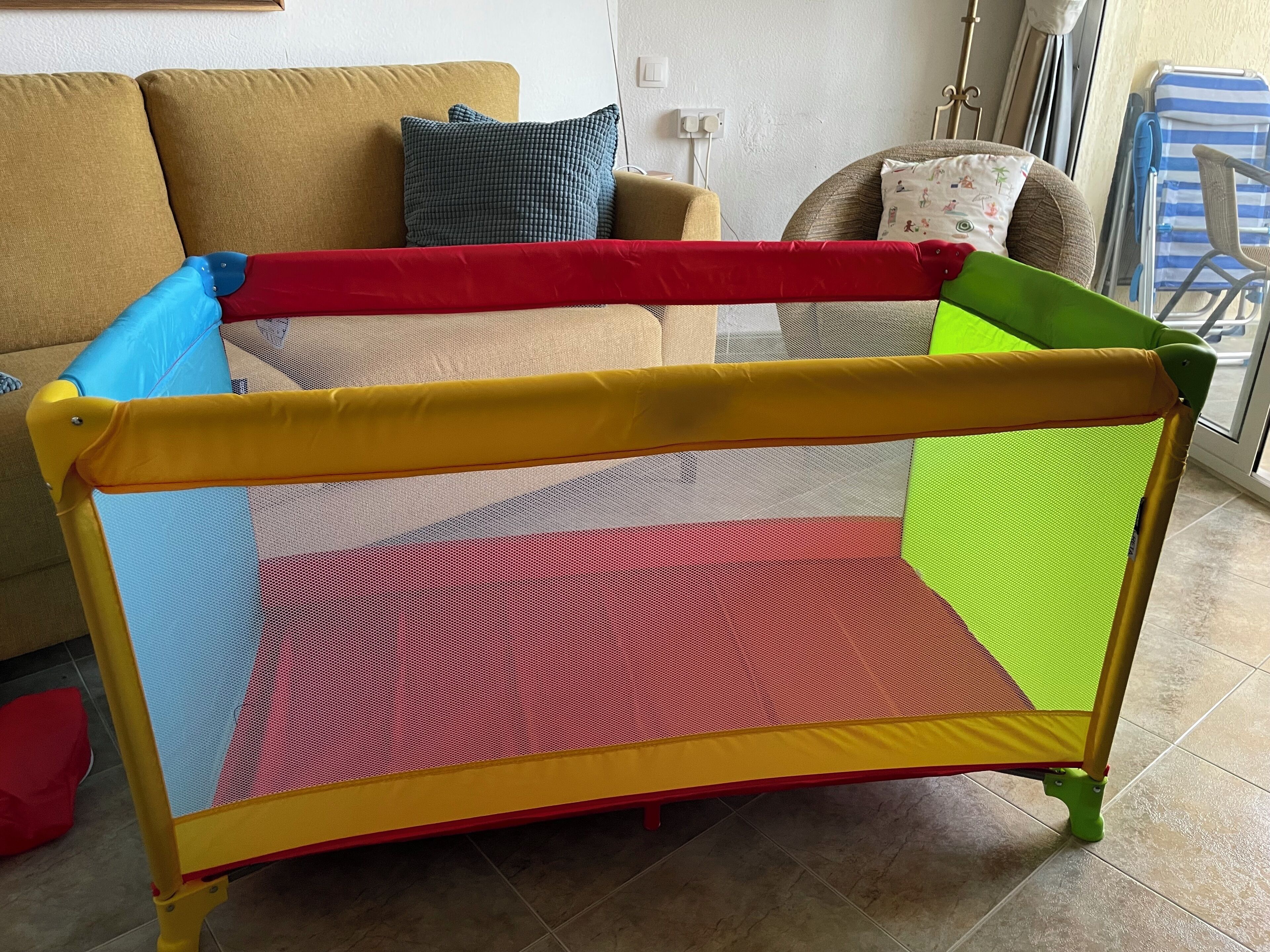 Ein Reisebett mit gelbem Gestell und Netzseiten, platziert in einem Wohnzimmer mit einem beigefarbenen Sofa und einem blauen Kissen.