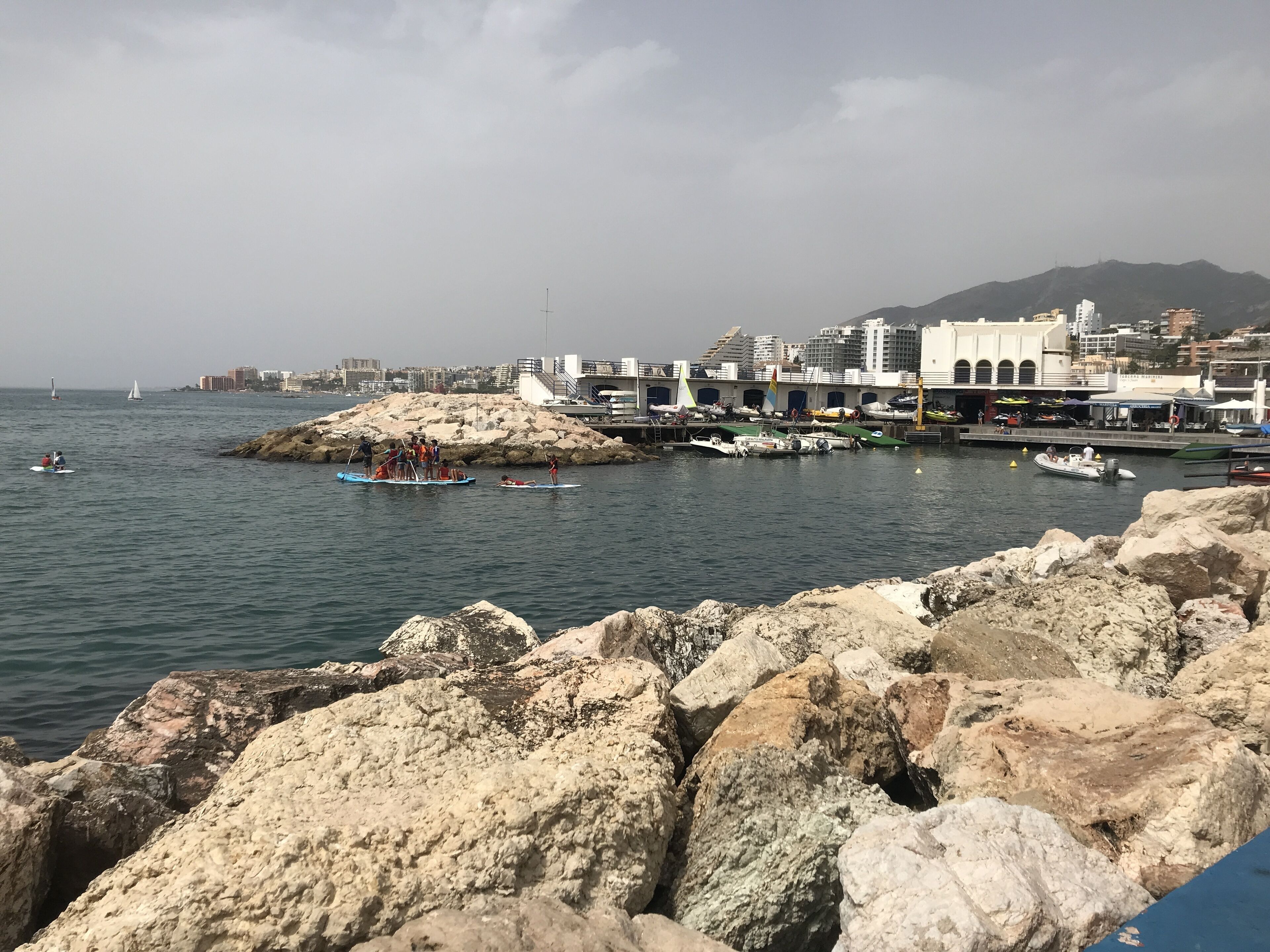 Eine Küstenlandschaft mit felsigem Gelände, einer Marina mit Booten und Gebäuden im Hintergrund.