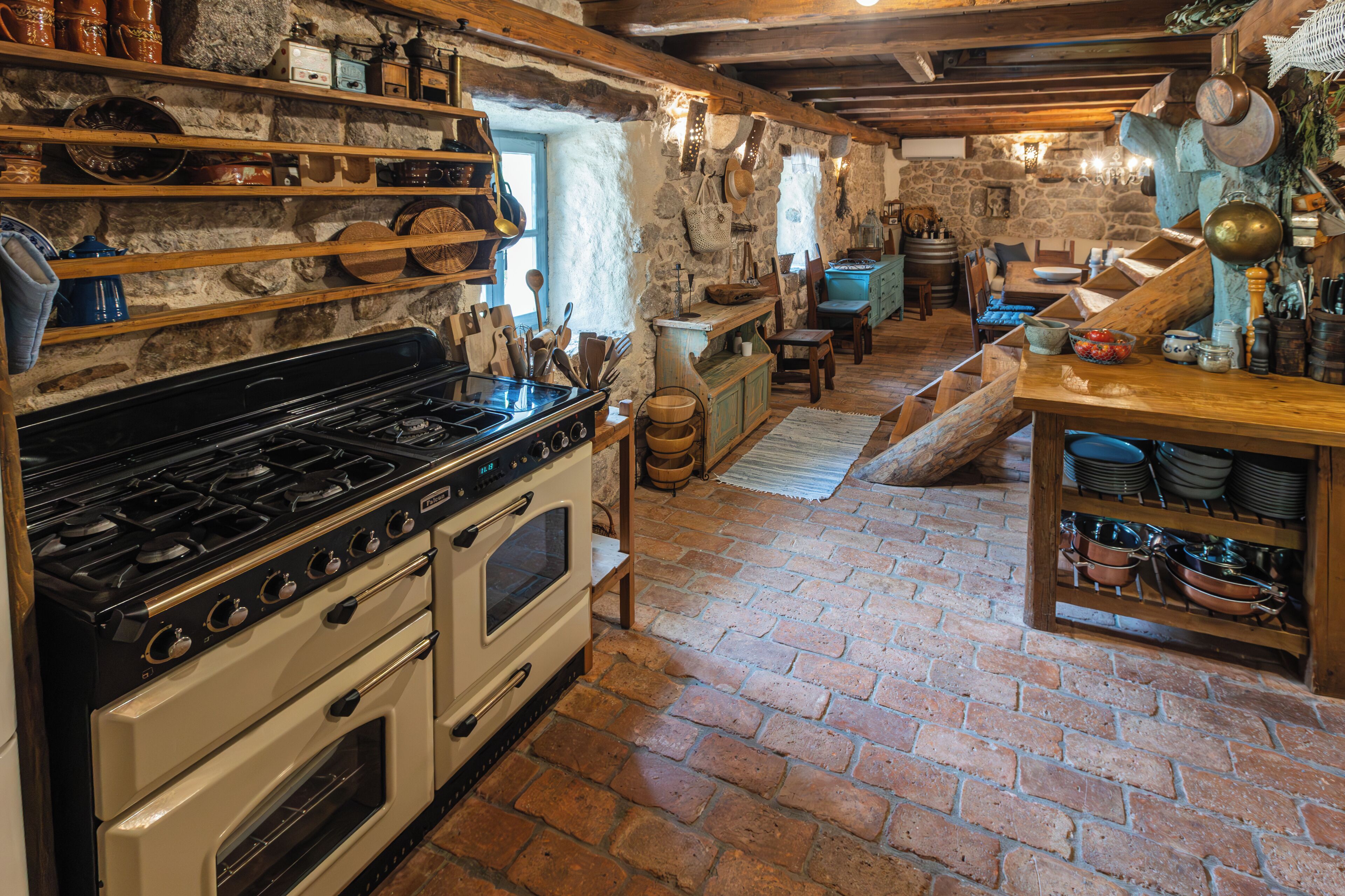 A rustic kitchen with a stove, oven, shelves with dishes, and a wooden table.