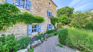 Un edificio in pietra con persiane blu, un vialetto ghiaioso e vegetazione rigogliosa.
