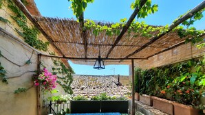 Een overdekt terras met een tafel en stoelen, omringd door potplanten en een uitzicht op een betegeld dak op de achtergrond.