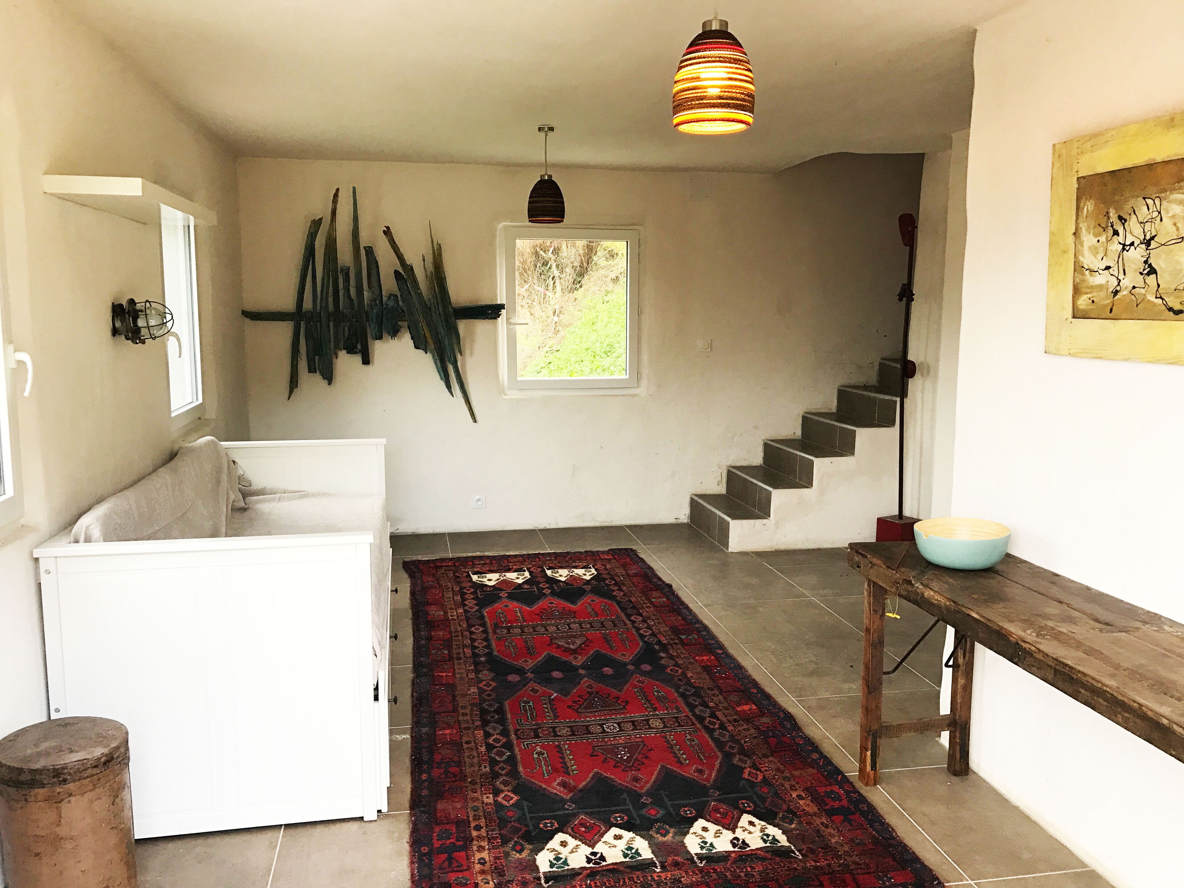 A room with a white sofa, a patterned rug, a wooden table, a bowl, a staircase, and a hanging light fixture.