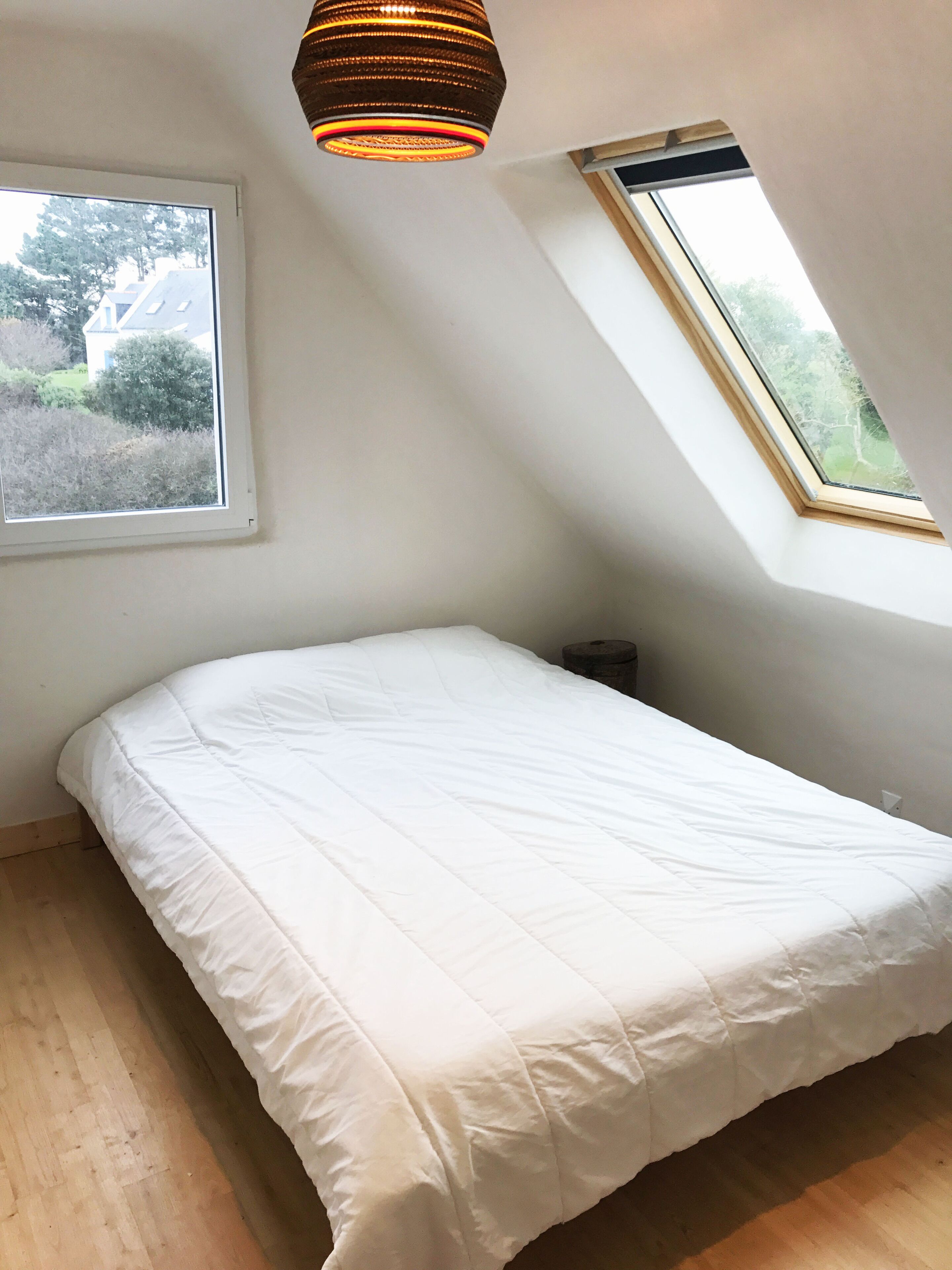 A bedroom with a sloped ceiling, a skylight, a bed with white bedding, and a hanging light fixture.