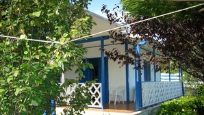 Un área de estar al aire libre con techo, valla blanca y detalles azules, rodeada de abundante vegetación.
