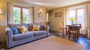 A living room with a sofa, chairs, a dining table, and a view of greenery through the windows. - Tin mine riverside retreat, log fires, views, wildlife, ruins, pet-friendly (Calstock)