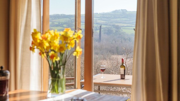 A table with a vase of yellow flowers, a wine bottle, and a glass of red wine, overlooking a scenic landscape with a distant tower. - Tin mine riverside retreat, log fires, views, wildlife, ruins, pet-friendly (Calstock)