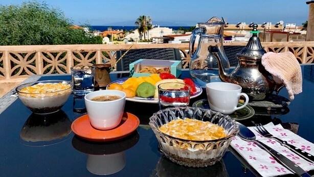 Mesa de pequeno-almoço com vários pratos, um bule e vista para o mar e edifícios.