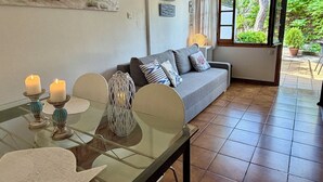 Ein Wohnzimmer mit Glastisch, zwei Kerzen, einer Couch mit Kissen, einem Bild von Strandhütten und einem Fenster mit Blick auf Grünflächen.