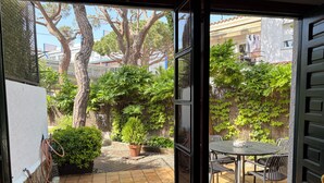 Eine geöffnete Schiebetür führt auf eine Terrasse mit Tisch und Stühlen, umgeben von Blumentöpfen und Bäumen.