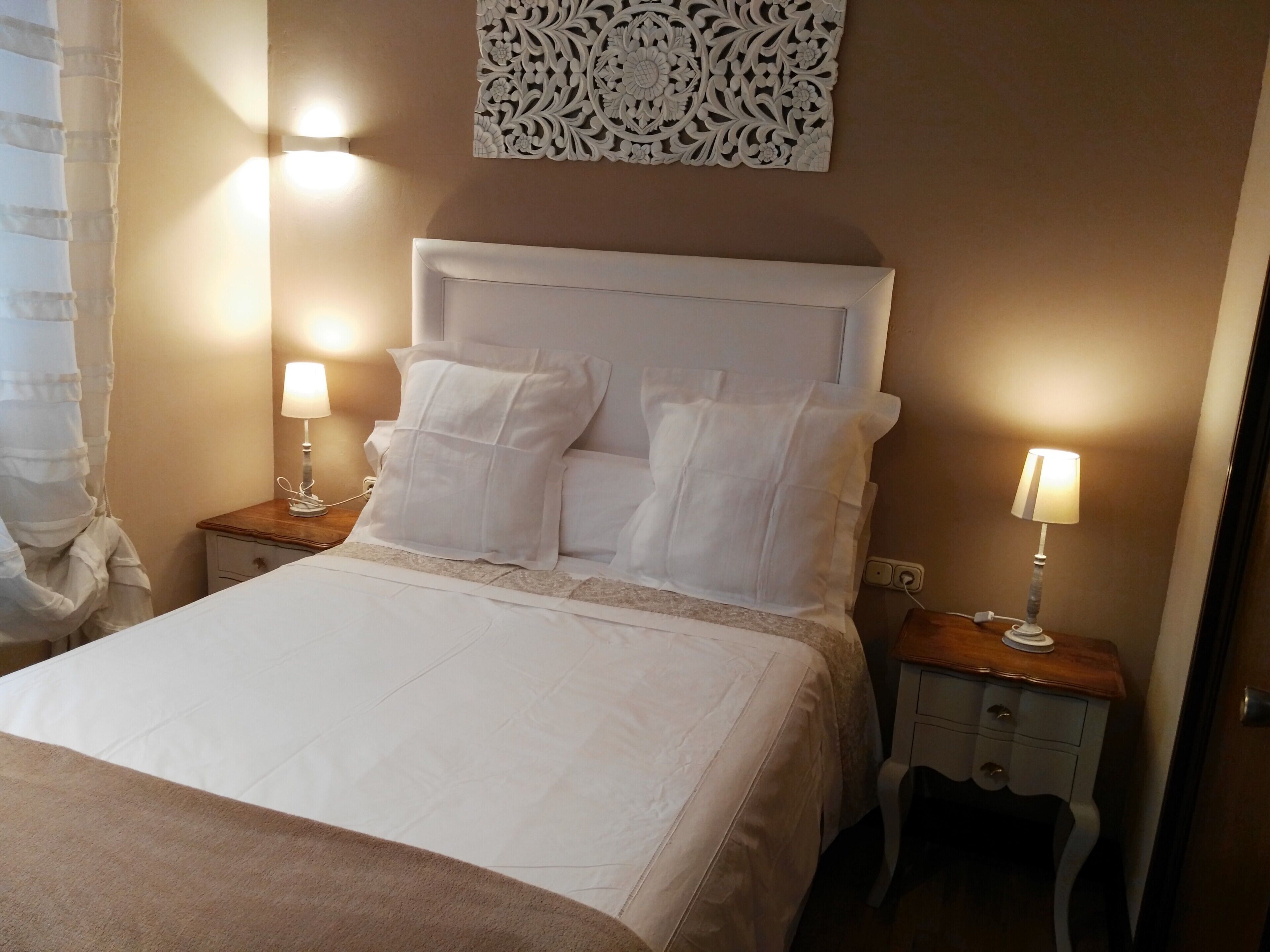 Una cama bien tendida con sábanas y almohadas blancas, dos lámparas de noche, un adorno en la pared y una mesita de noche de madera con un cajón.