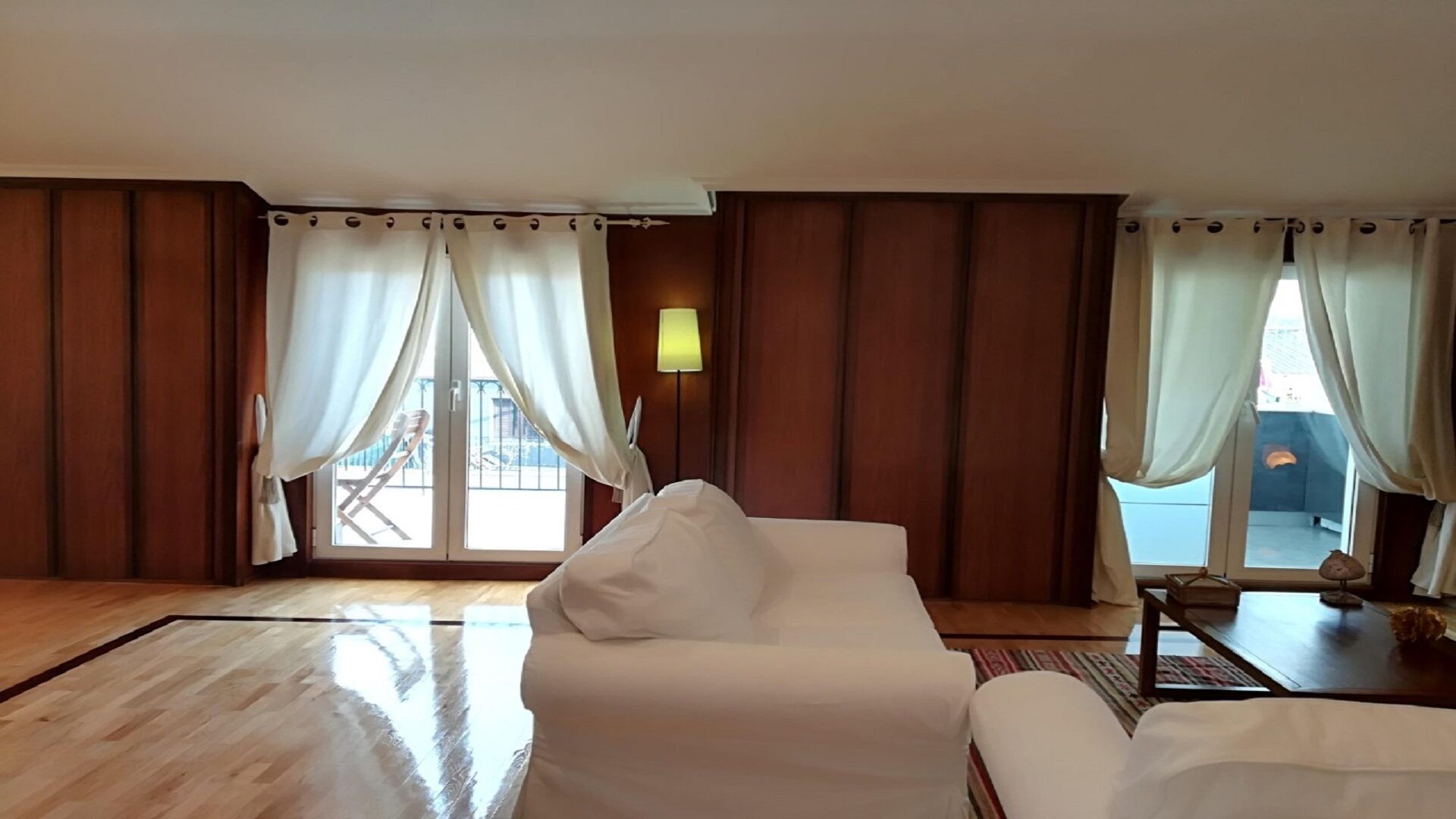 A hotel room with a large bed, a white armchair, a small table with a lamp, and a view of the outside balcony through sheer curtains.
