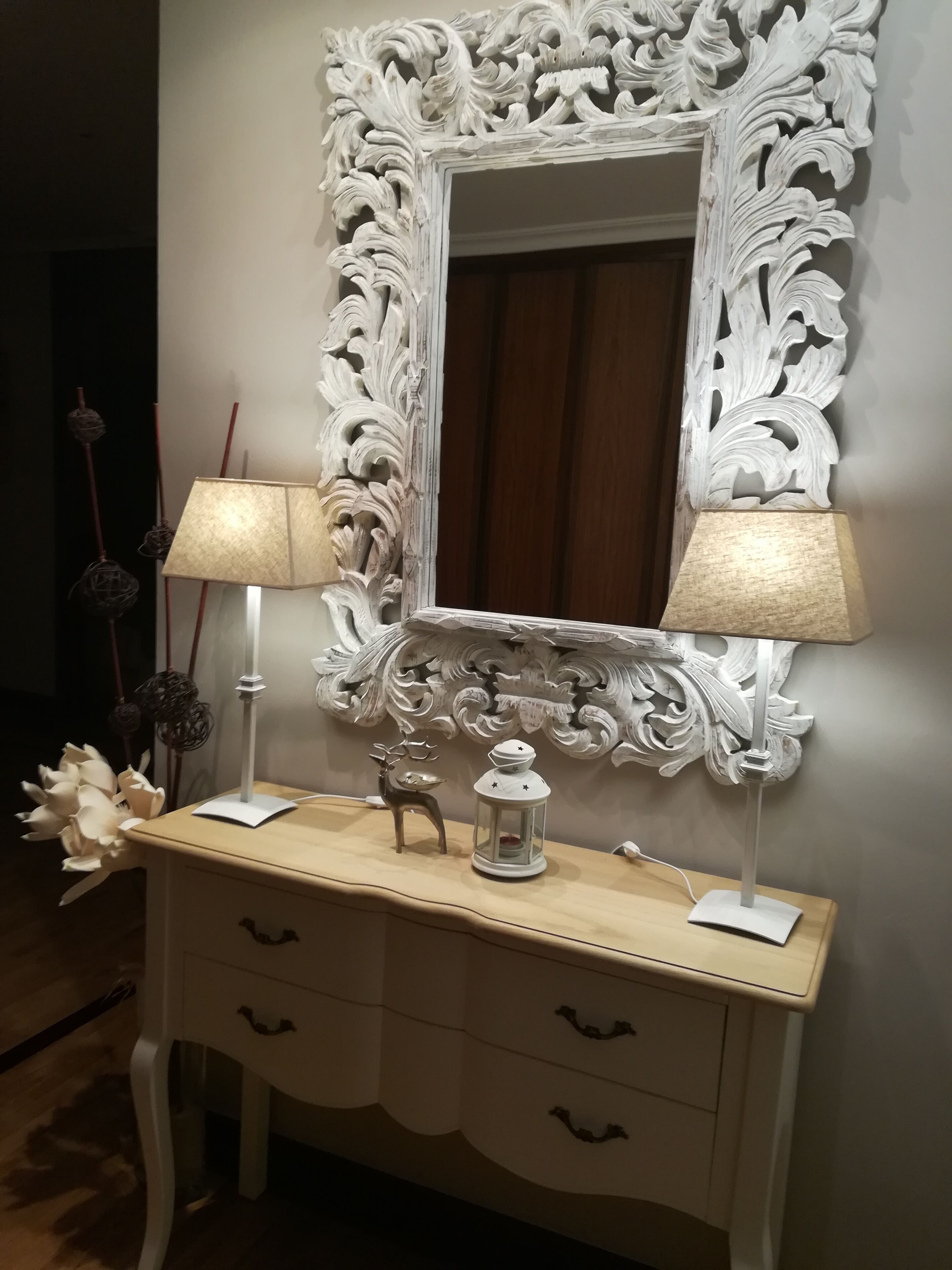 Un espejo blanco ornamentado con un marco decorativo, una consola de madera con cajones, dos lámparas, un pequeño ciervo decorativo y un farol.