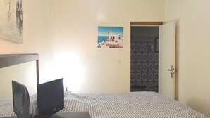 Un dormitorio con cama, escritorio con computadora, puerta y pared con un cuadro.