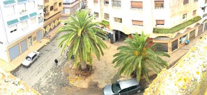 Vista de la calle con tiendas frente a edificios residenciales, palmeras y coches aparcados.
