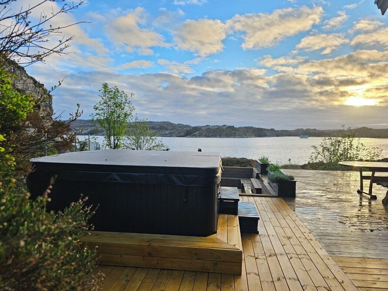 A wooden deck with a hot tub, outdoor seating, and a view of a lake at sunset.
