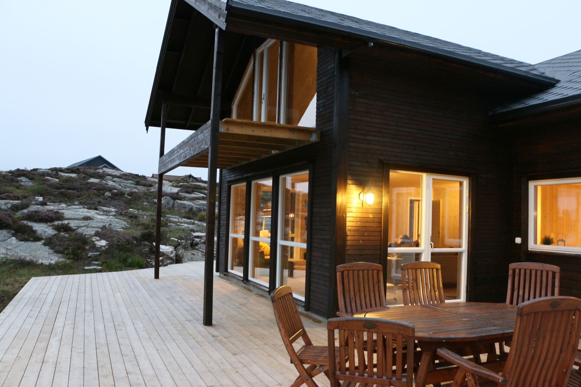 A modern wooden cabin with a deck, large windows, and a dining area with chairs.