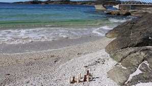 A pebble beach with clear water, a rocky shoreline, and a bridge in the background.