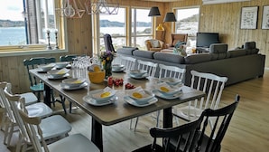 A dining area with a wooden table set for a meal, a sofa, and a television in a room with wooden walls and large windows overlooking a body of water.