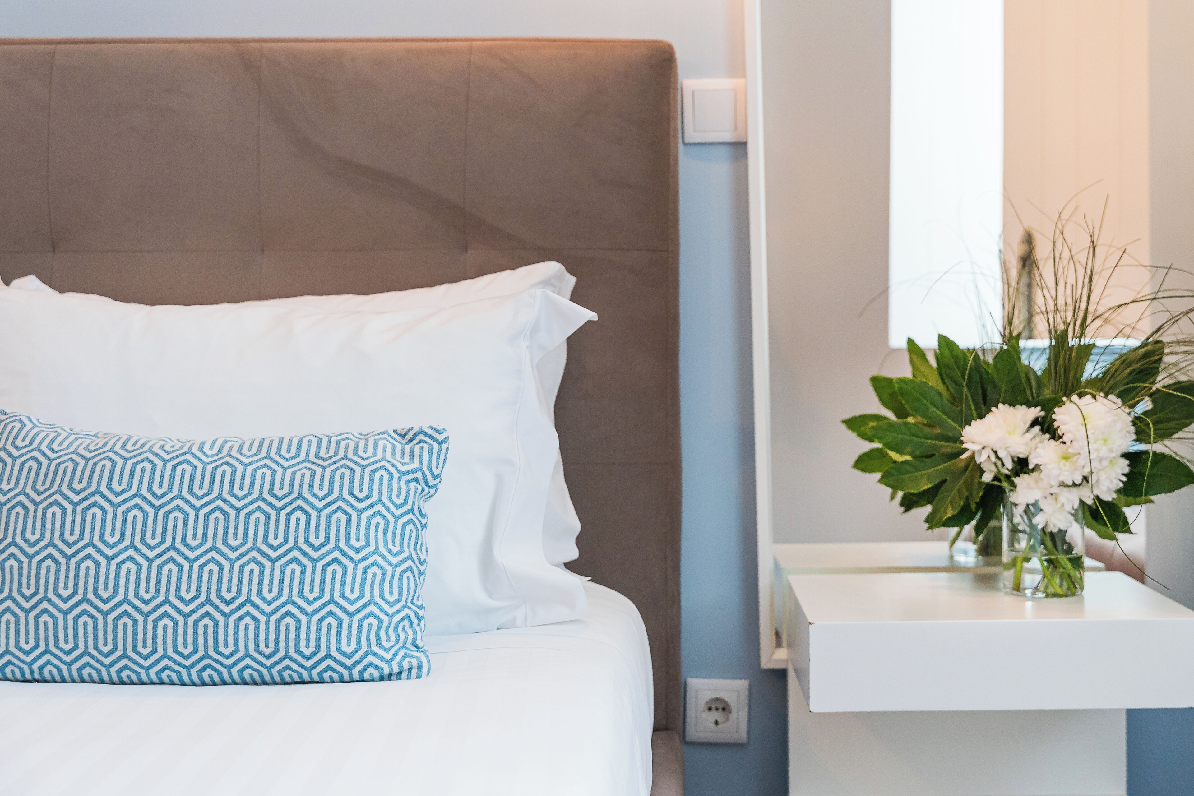 A bed with white and blue patterned pillows, a brown headboard, and a white bedside table with a vase of white flowers.