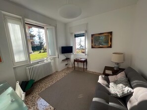 Uma sala de estar com sofá, televisão, pintura e janela com vista para um jardim.