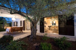 An outdoor seating area with a wooden bench and table, surrounded by greenery and a large tree.
