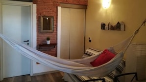 Een kamer met een hangmat, een bakstenen muur, een deur, een kleine tafel met een plant, en een wandplank met decoratieve objecten.