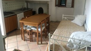 Una cocina compacta con una mesa y sillas, una cama con estructura metálica, y un ventanal con contraventanas.