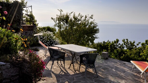 Un patio al aire libre con una mesa y sillas de piedra, rodeado de abundante vegetación y vistas al mar.