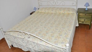 Una cama con colcha floral, una mesita de noche con lámpara y un aparador de madera con cajones.