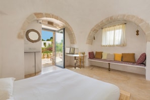 A bedroom with a bed, a desk, and a bench with cushions. There are stone arches and a view of the outdoors.