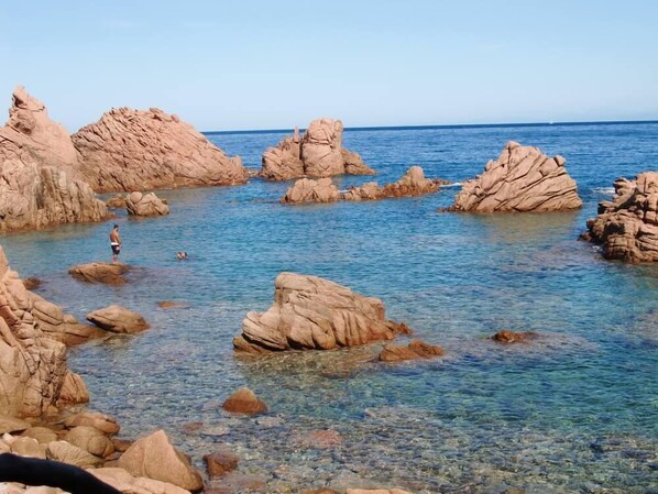 Una zona costera rocosa con agua cristalina y una persona de pie sobre una roca.