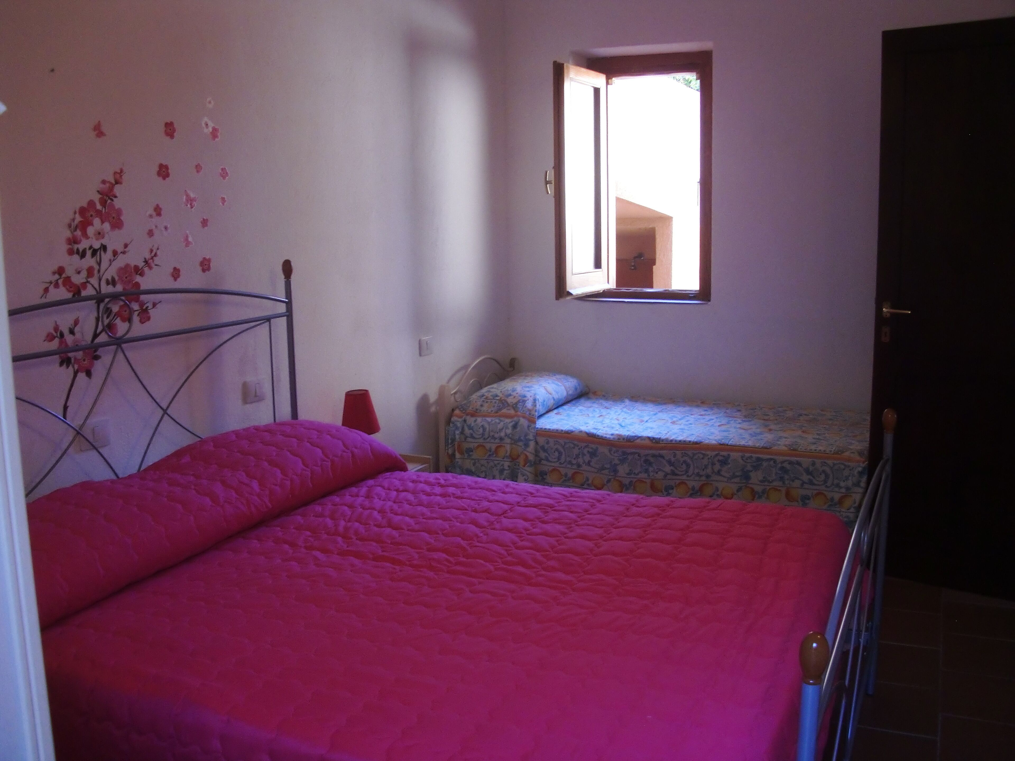 A bedroom with two beds, one with a pink bedspread and the other with a patterned bedspread. There is a window with a view of the outside, a door, and a wall with a floral decal.