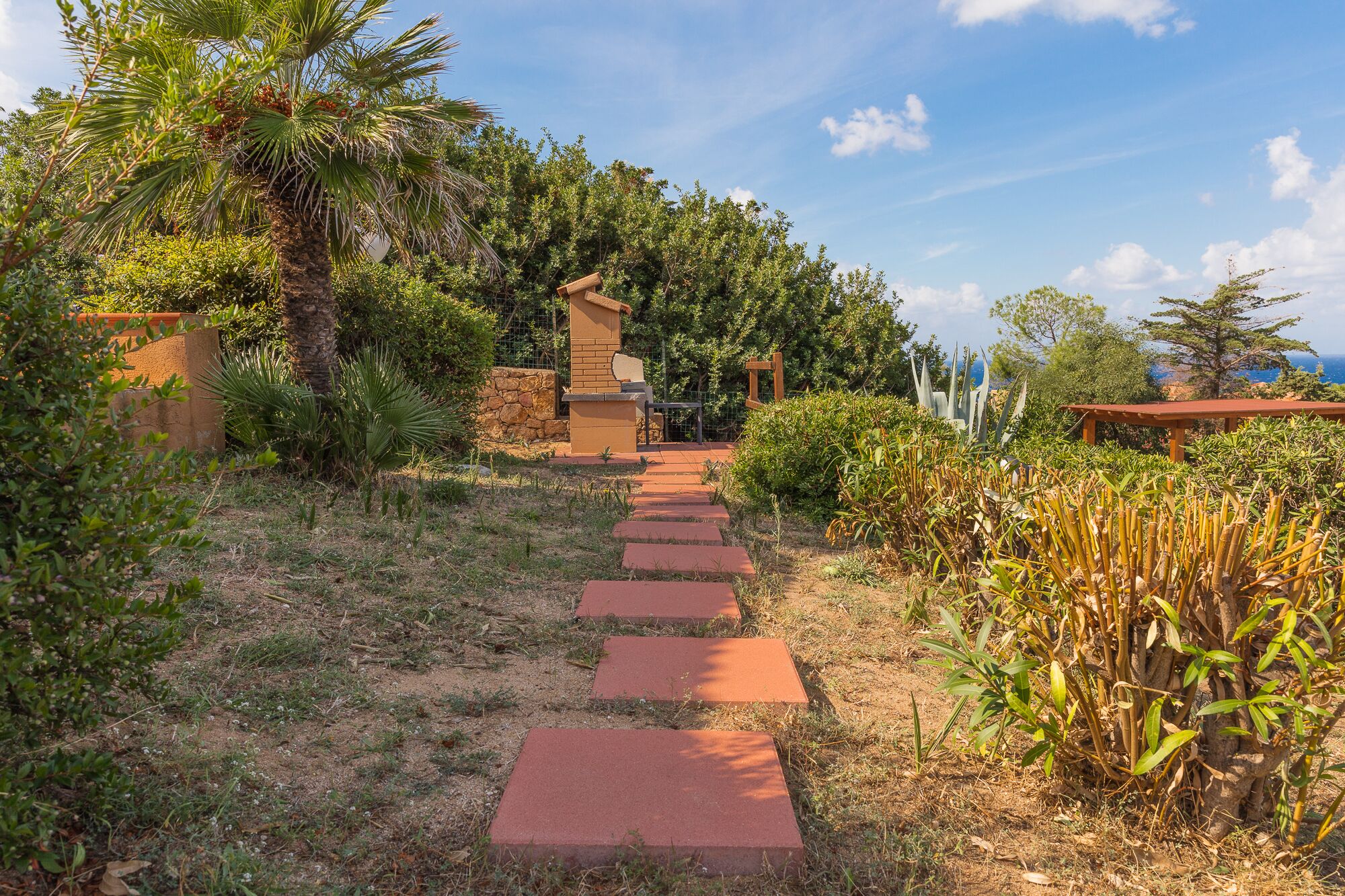 A garden pathway leading to a wooden structure, surrounded by various plants and a clear sky.