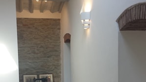 Un pasillo estrecho con una cama a la izquierda, un muro de piedra, una pequeña mesa con una máquina de coser y una alfombra roja.