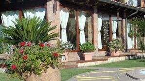 A large planter with various plants and flowers in front of a building with brick walls and large windows. - Villa Graziosa (Quartu sant'Elena)