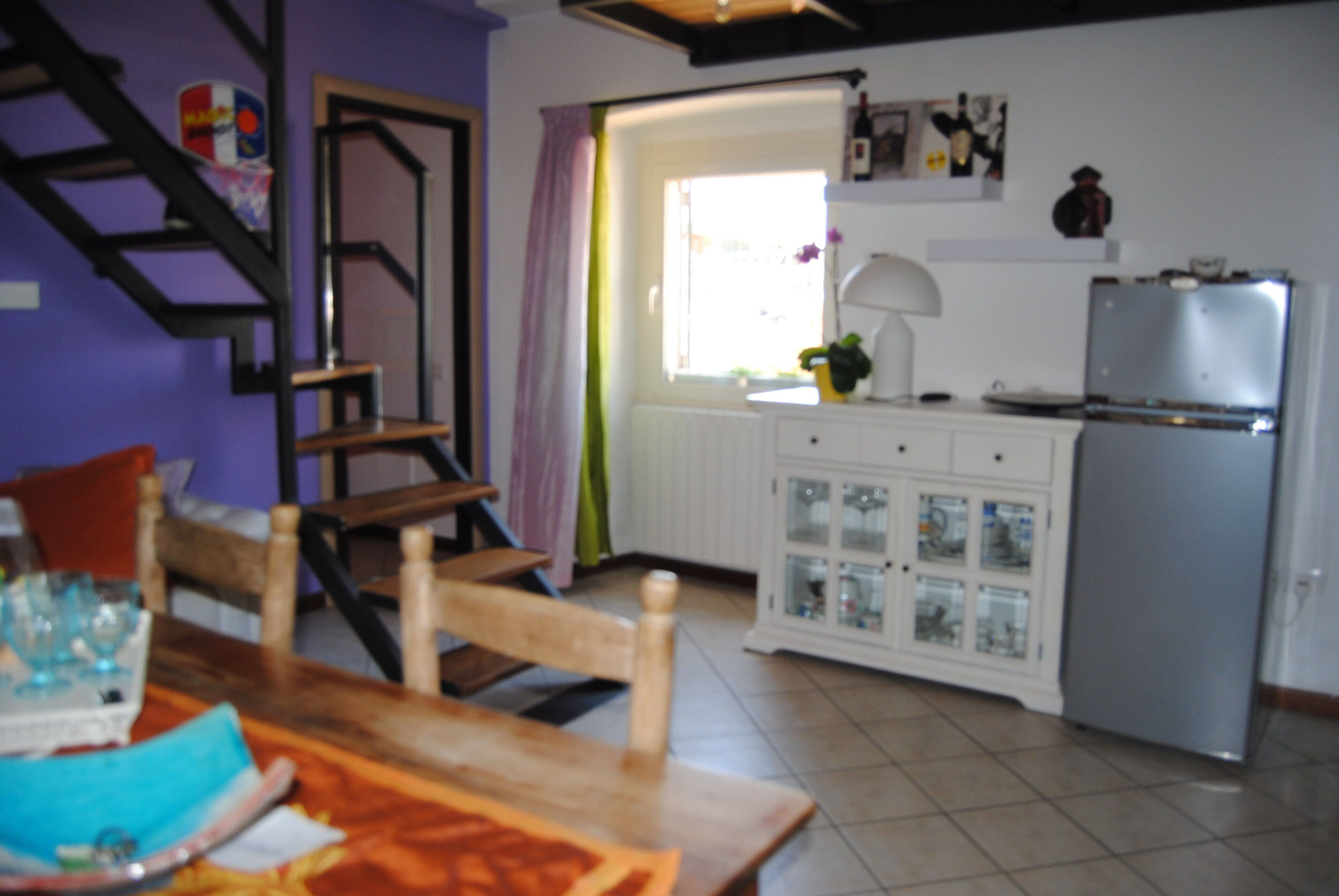 Una cucina con frigorifero, una finestra con tende, un pensile con ante in vetro e un tavolo in legno.