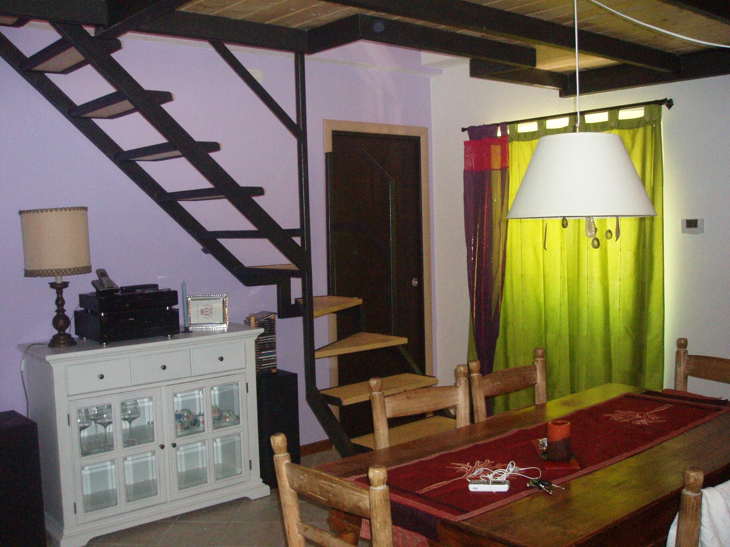 Zona pranzo con tavolo in legno, sedie, runner rosso, credenza bianca con ante in vetro, lampada e una scala.