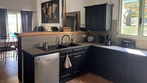 A compact kitchen with dark cabinetry, a built-in dishwasher, a sink, and a window overlooking a garden. - House / Villa - Sainte sabine (Beaumontois En Périgord)