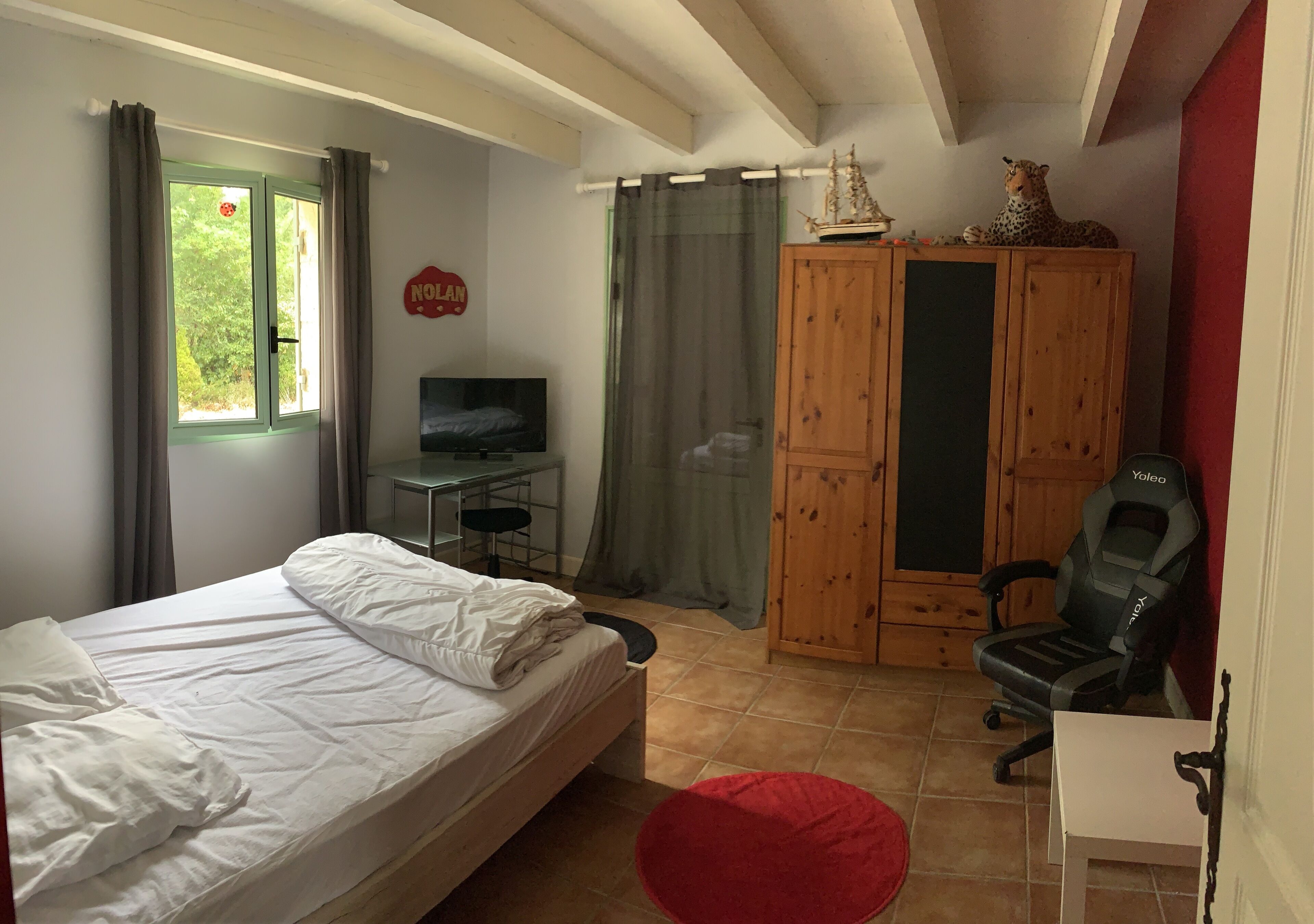 A bedroom with a bed, a TV, a wooden wardrobe, a desk, and a red ottoman.