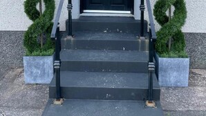 Una puerta frontal negra con un letrero con el número '6', una pequeña ventana con diseño de rombos y una escalera de metal negra con barandilla.
