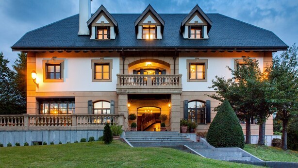 Une grande maison à deux étages, dotée d’une façade symétrique, d’une entrée centrale avec un balcon et d’une pelouse bien entretenue.