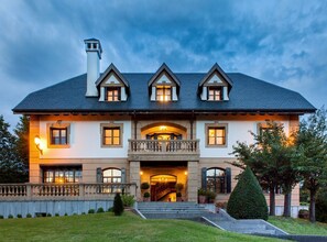 Une grande maison à deux étages, dotée d’une façade symétrique, d’une entrée centrale avec un balcon et d’une pelouse bien entretenue.