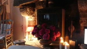 Ein gemütlicher Raum mit Kamin aus Stein, einem Holzstuhl, einem kleinen Tisch mit Kerzen und einem Blumenstrauß.