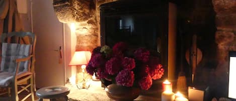 Une pièce chaleureuse avec une cheminée en pierre, une chaise en bois, une petite table avec des bougies et un vase rempli de fleurs.