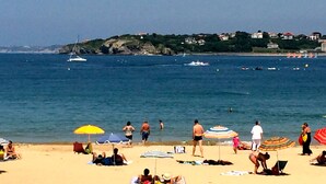 Uma praia com pessoas tomando sol e nadando, um veleiro e casas ao fundo.