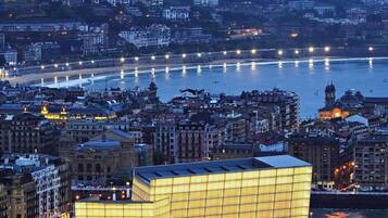 Un paisaje urbano costero al anochecer con un edificio moderno destacado de ventanas iluminadas, una playa y un río con barcos.