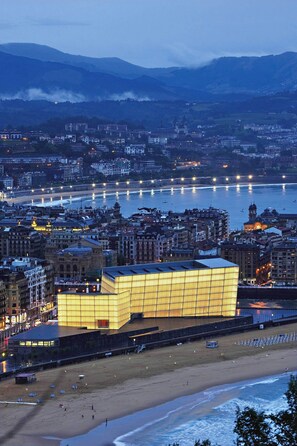 Een kuststadsgezicht in de schemering met een opvallend modern gebouw met verlichte ramen, een strand en een rivier met boten.