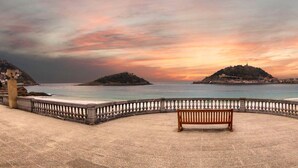 Une promenade en bord de mer agrémentée d’un banc, d’une balustrade en pierre et offrant une vue sur la mer et les collines lointaines.